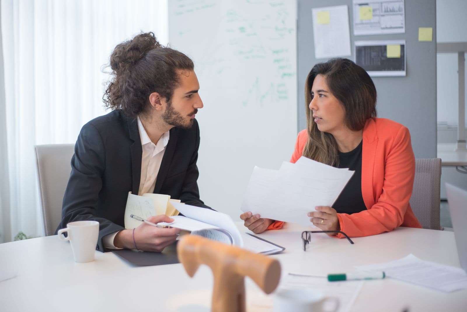 Deux collègues en discussion à une table de bureau, avec des documents et un café.